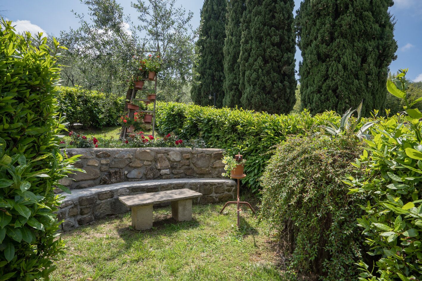 A stone bench in a lush garden with a variety of plants and a tiered planter with flowers.