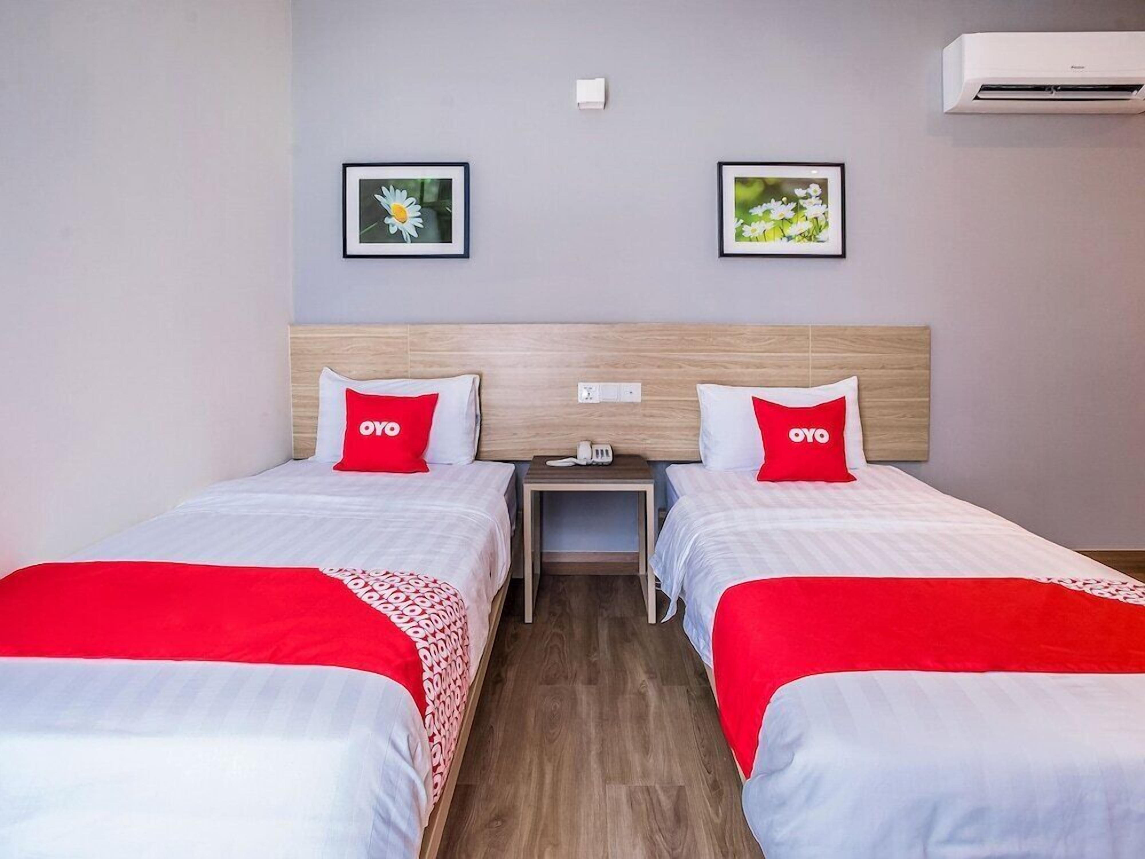 A hotel room with two beds, each with a red OYO pillow, a nightstand, a wall-mounted air conditioner, and two framed pictures on the wall.