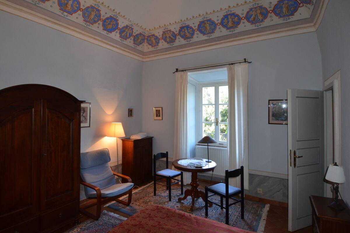 Een kamer met een houten kast, twee stoelen, een tafeltje met een lamp, een raam met gordijnen, en een deur.