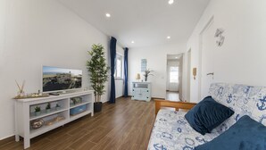 Una sala de estar moderna con piso de madera, una unidad blanca para entretenimiento, un sofá azul con almohadas de temática náutica y un televisor sobre un pedestal.