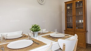 Un comedor con una mesa blanca preparada para cuatro personas, con platos, cubiertos y copas, un reloj en la pared y un armario de madera al fondo.