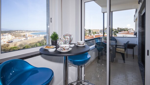 A balcony with a table set for tea, blue bar stools, and a view of the beach and city. - B17 - The Stunning Seaview Apartment (Lagos)