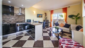 A modern kitchen with a central island, a dining area with a sofa and chairs, and a living room with a large window and curtains. - B47 - Walkway Beachview Place (Lagos)