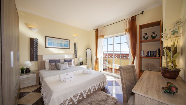 A hotel room with a bed, a desk, a chair, a bookshelf, and a balcony with curtains. - B47 - Walkway Beachview Place (Lagos)
