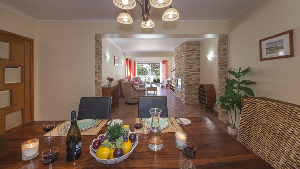 A dining area with a wooden table set for a meal, a chandelier, and a view into a living room with a stone wall and a plant. - D07 - Torraltinha Villa in Lagos (Lagos)