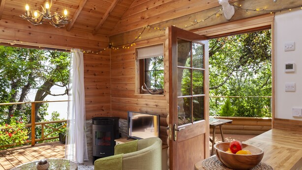 A wooden cabin interior with a dining table set for two, a fireplace, and a large window overlooking greenery. - Porthole Log Cabin (Minehead)