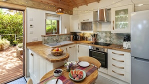 A modern kitchen with wooden cabinets, a stainless steel range hood, and a built-in oven. There is a breakfast bar with a fruit bowl and a vase of flowers. - Porthole Log Cabin (Minehead)