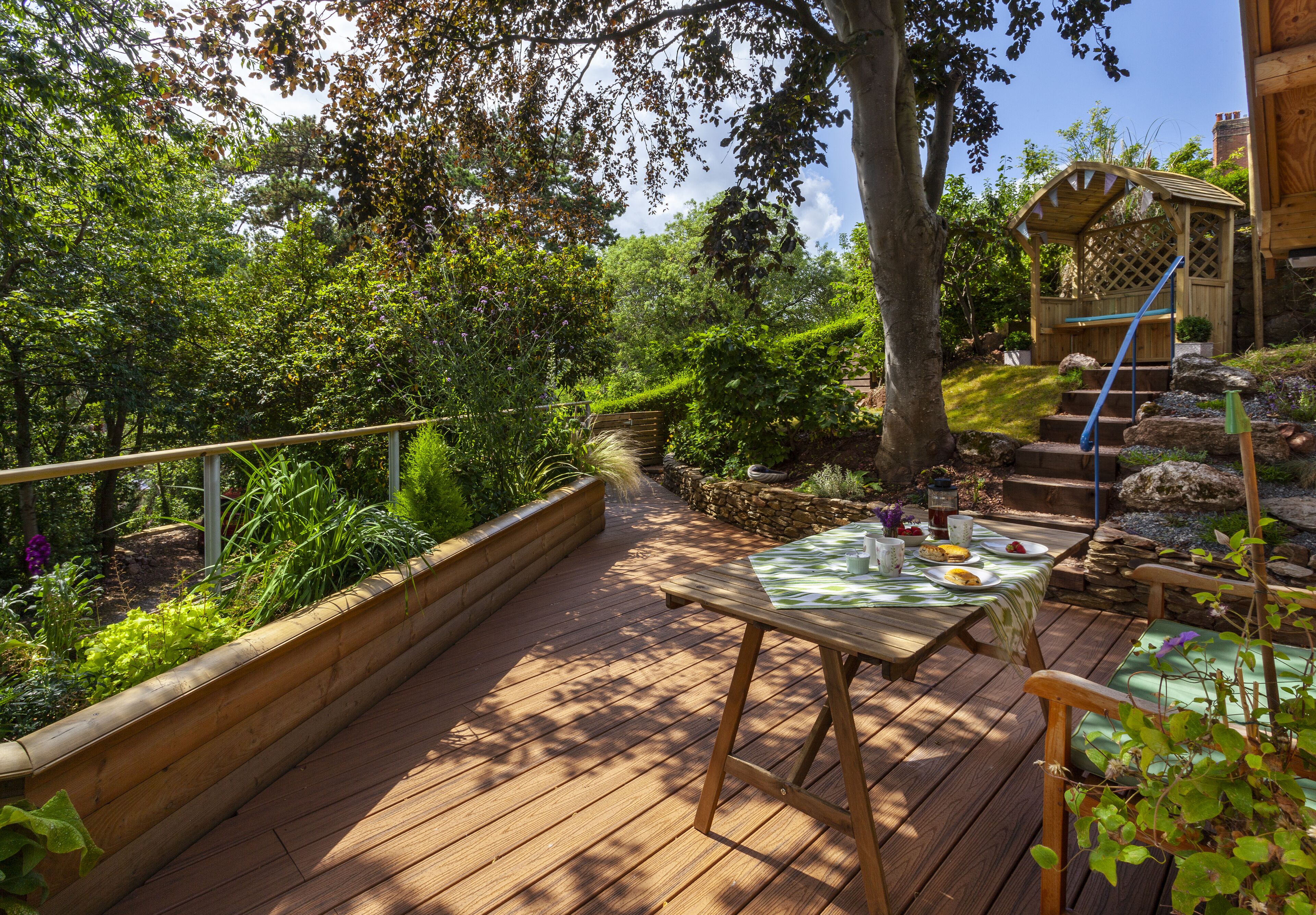 Eine Holzterrasse mit Tisch und Stühlen, umgeben von üppiger Vegetation und einer Holzpergola.