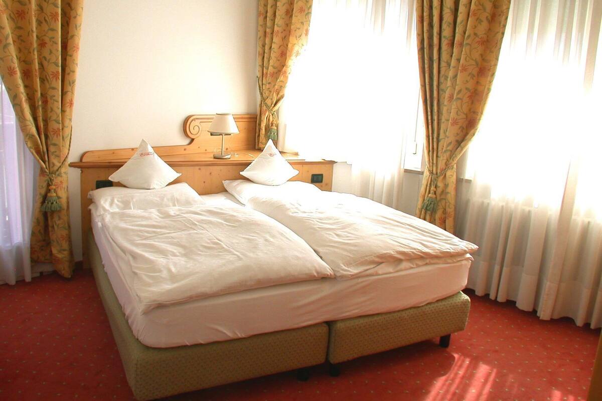 A double bed with white linens, a wooden nightstand with a lamp, and floral curtains.