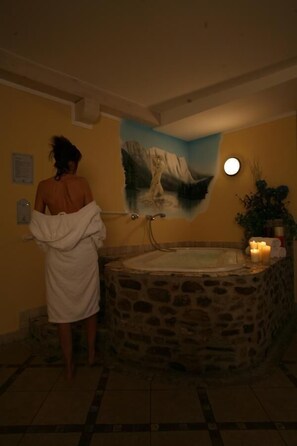A person in a white bathrobe standing by a stone bathtub with a painting of a mountain scene above it. - Hotel Eccher (Mezzana)