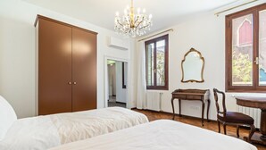 A bedroom with two beds, a wooden wardrobe, a desk, a chair, a mirror, and a chandelier. - GuestHost - Large and bright 145sqm apartment with terrace, located in the center of Venice (sestriere Castello), 5 mins from San Marco and Rialto Bridge and 2mins from the lively Campo S.Maria Formosa. Located on the second floor, without lift, of a Ven (Venice)