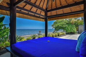 A room with a blue bed, a view of the sea, and a thatched roof structure.