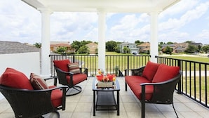 Un patio cubierto con mobiliario de mimbre y cojines rojos, una mesa pequeña con fruta, y vistas a un barrio residencial.