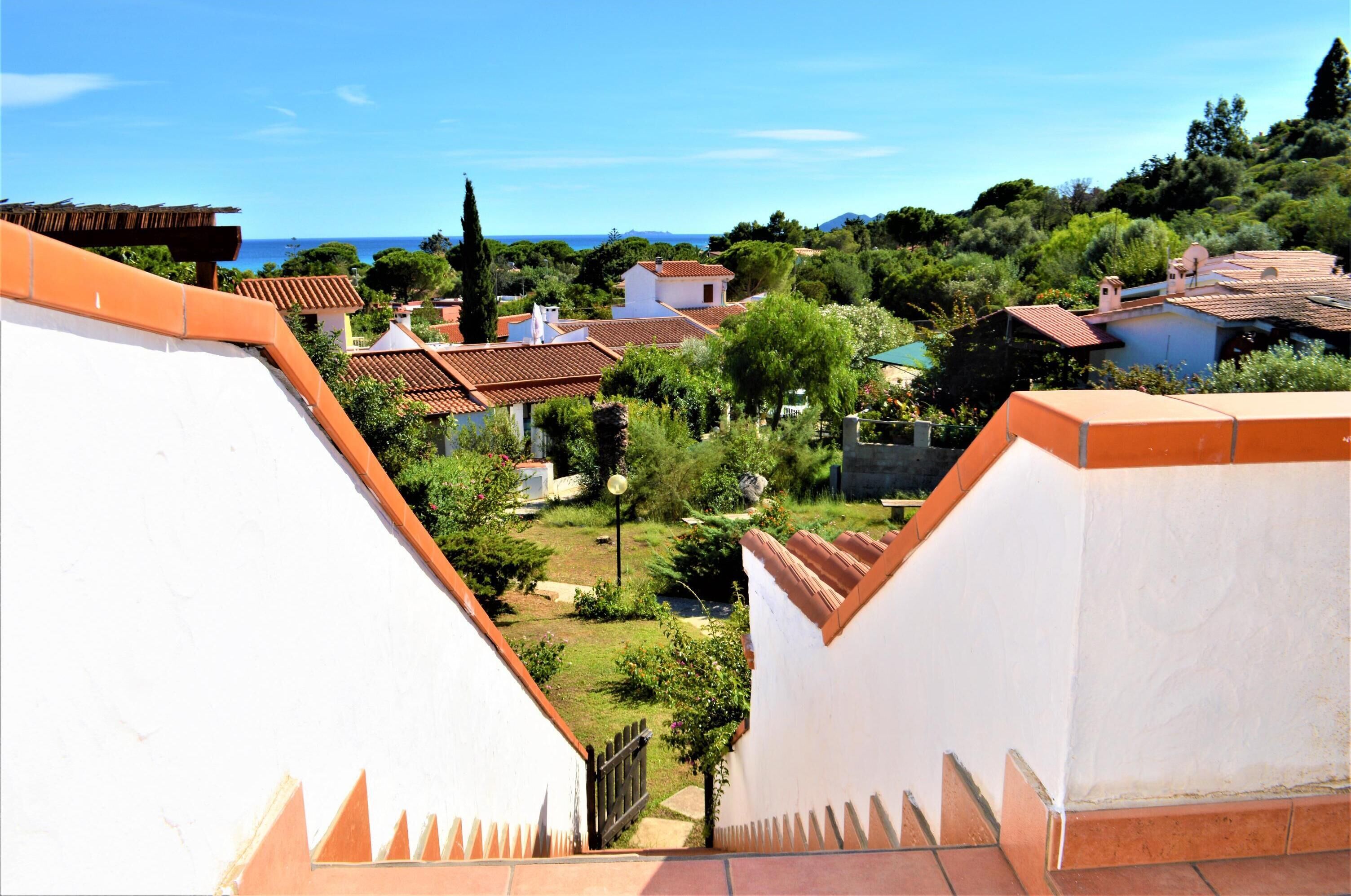 A residential area with terracotta-roofed houses, a clear blue sky, and a view of the sea.