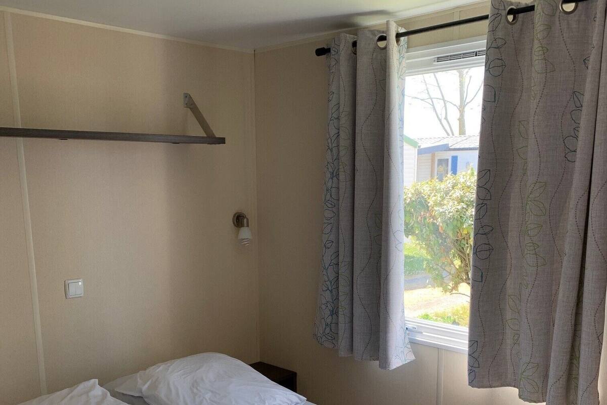A single bed with white linens, a brown towel, and a window with grey curtains.