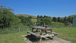A picnic table with a pot on top, surrounded by a grassy area with a fence and trees in the background. - The Little Bunk House (Oswestry)