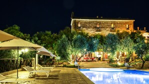 A nighttime pool area with lounge chairs and a large umbrella, surrounded by trees and a historic building in the background. - Masseria Mazzei Le Colline del Gelso (Corigliano-Rossano)