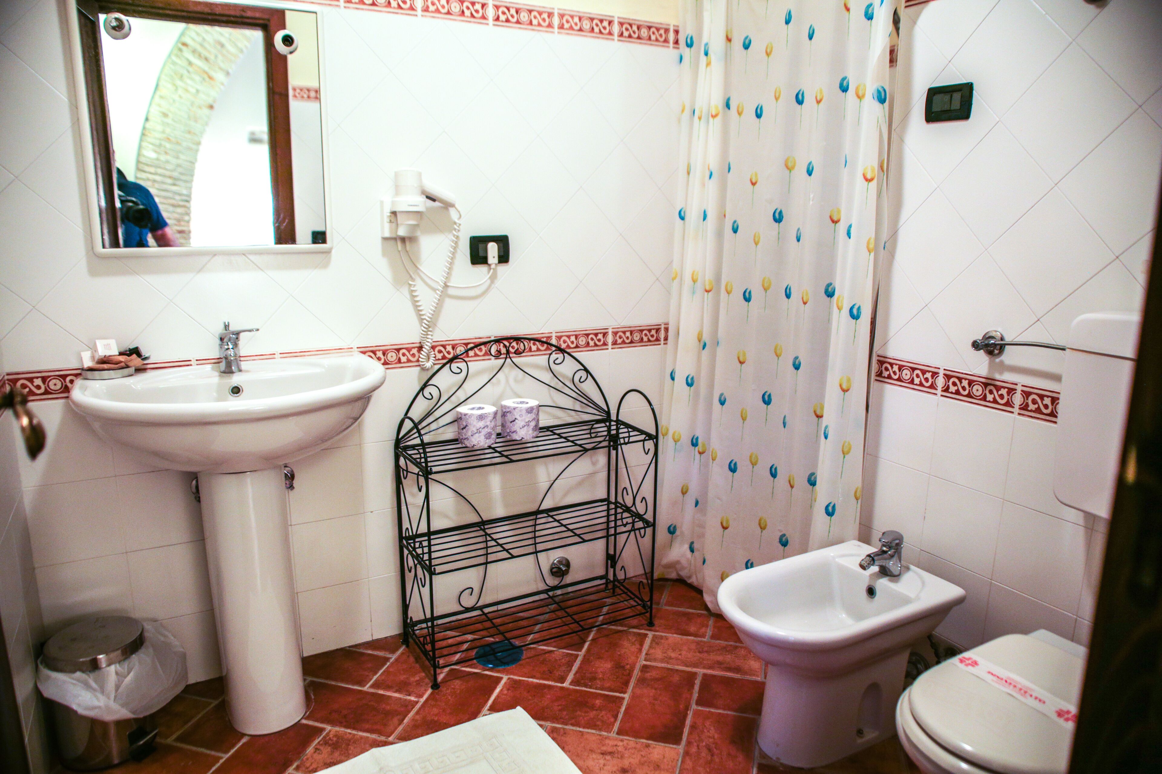 Ein Badezimmer mit Waschbecken, Bidet und Toilette. Über dem Waschbecken befindet sich ein Spiegel, und der Duschvorhang ist mit einem floralen Muster versehen.
