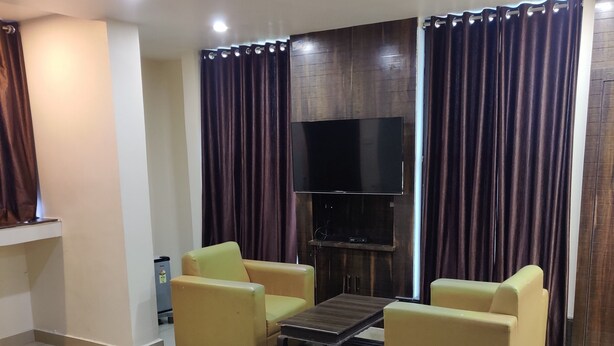 Une chambre d’hôtel avec un lit, deux fauteuils jaunes, une table basse, un téléviseur à écran plat fixé à un mur en bois, et une grande fenêtre avec des rideaux foncés.
