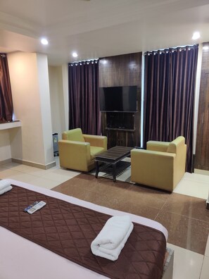 Une chambre d’hôtel avec un lit, deux fauteuils jaunes, une table basse, un téléviseur à écran plat fixé à un mur en bois, et une grande fenêtre avec des rideaux foncés.