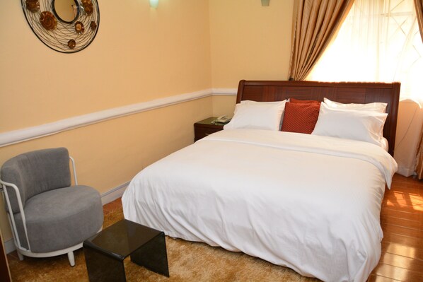 A neatly made bed with white linens, a gray armchair, and a small table in a room with beige walls and wooden flooring. - Aishar Residence (Lagos)