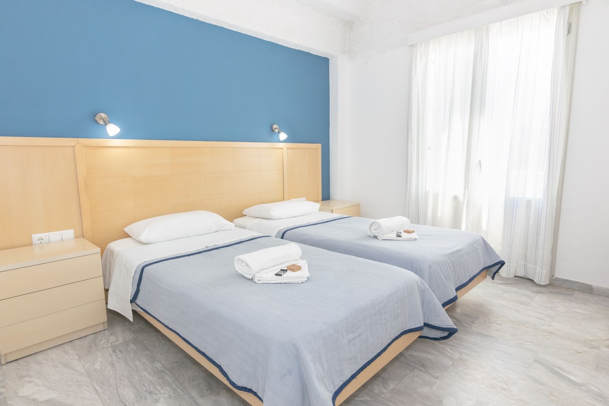 Quarto de hotel com duas camas, cabeceira de madeira, abajures e uma janela grande com cortinas.