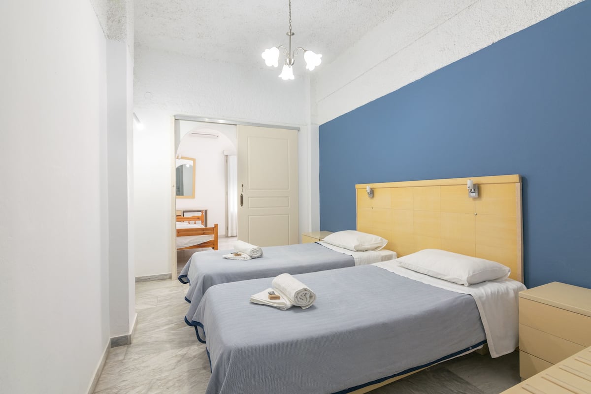 Quarto de hotel com duas camas, cabeceira de madeira, mesa de cabeceira e banheiro visível através de uma porta aberta.