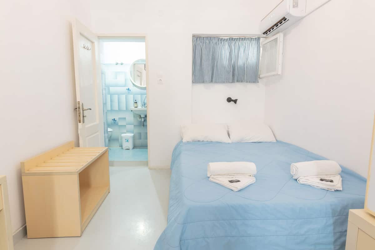 Quarto com cama, um banquinho de madeira, banheiro visível através de uma porta aberta e janela com cortinas.