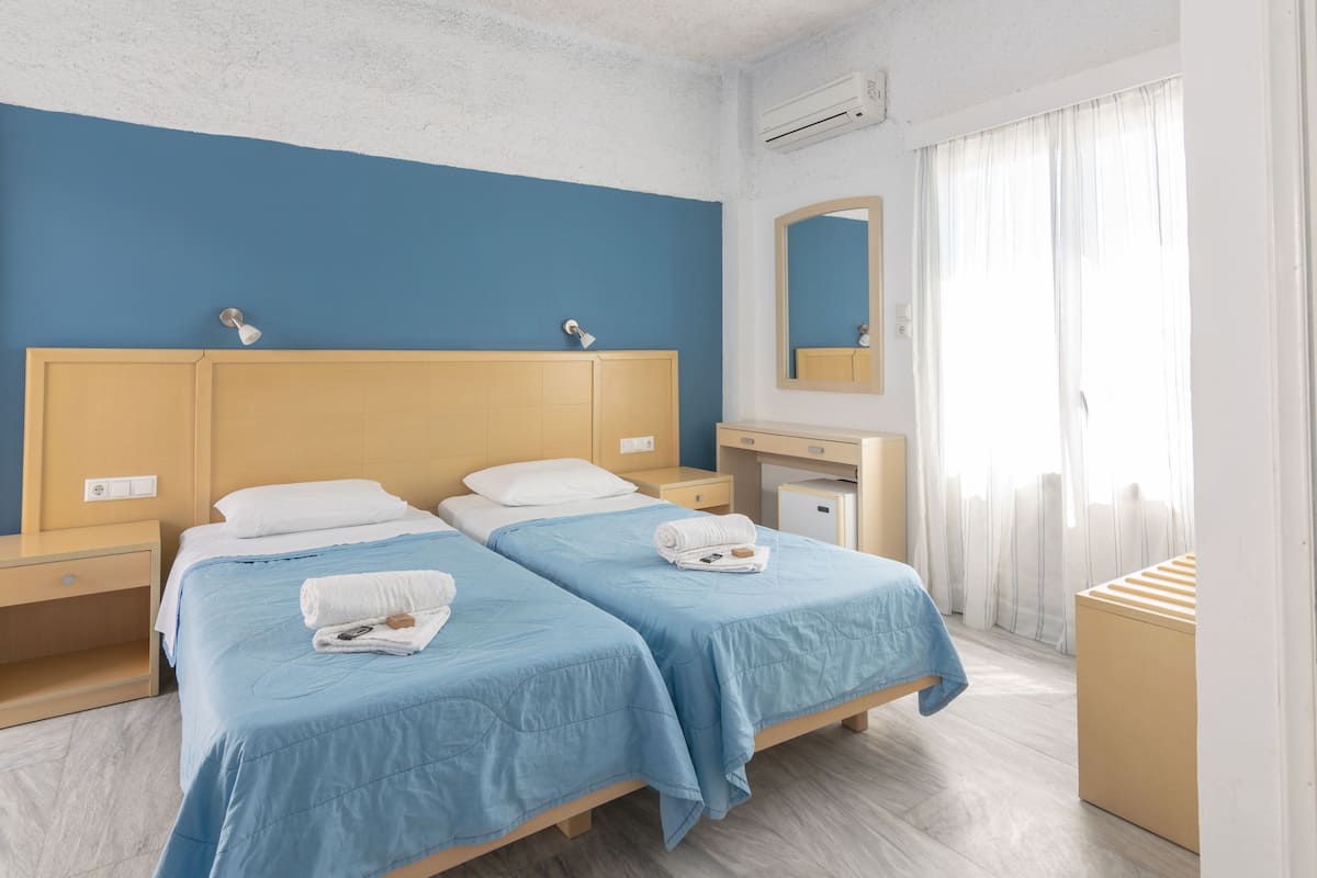 Quarto de hotel com duas camas, uma escrivaninha pequena, um espelho e uma janela com cortinas.