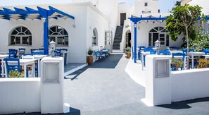 Un espacio al aire libre cubierto con mobiliario azul y blanco, un sendero central y edificios blancos con ventanas arqueadas.