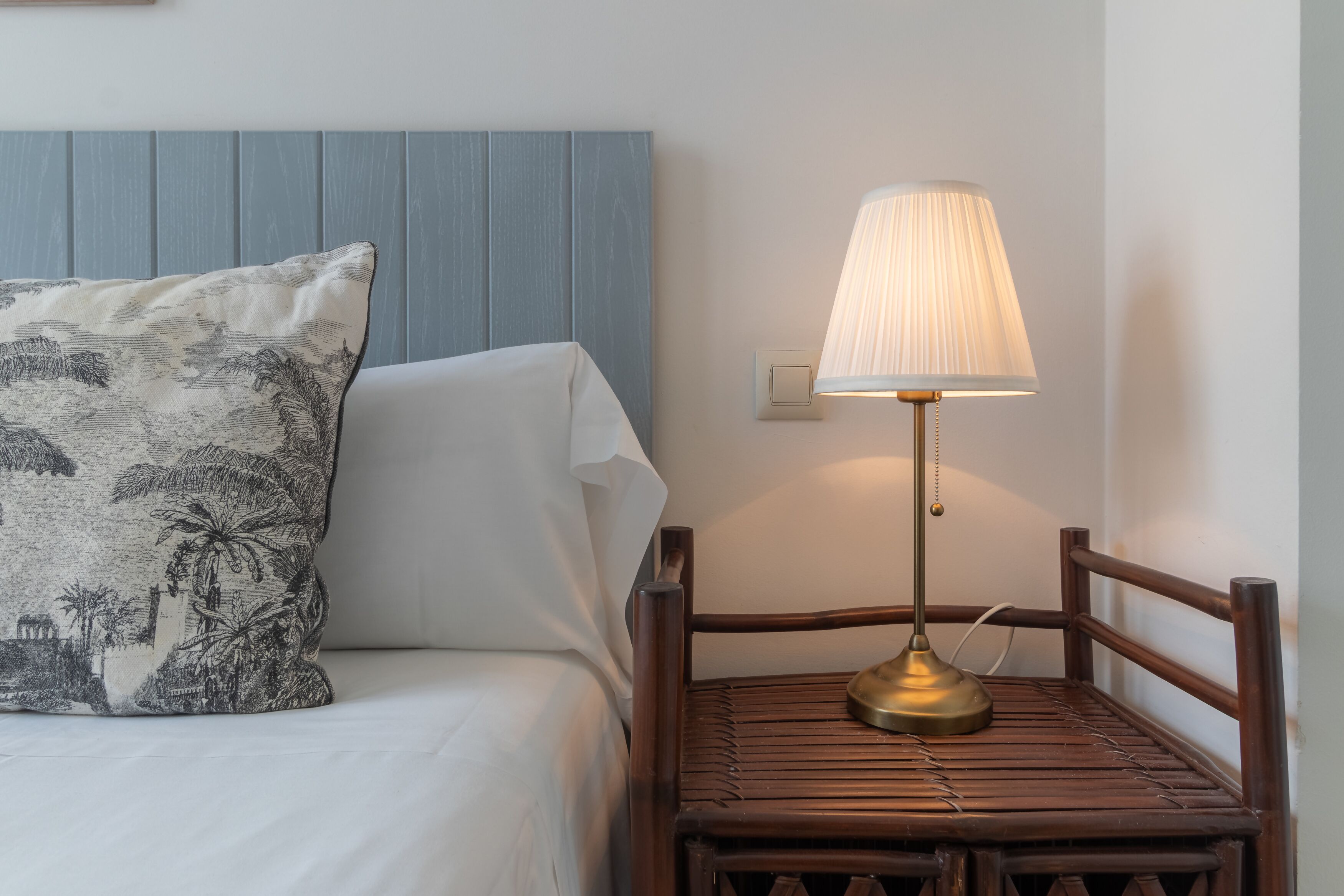 A bed with a white sheet and a decorative pillow, a wooden chair with a lamp, and a wall with a light switch.