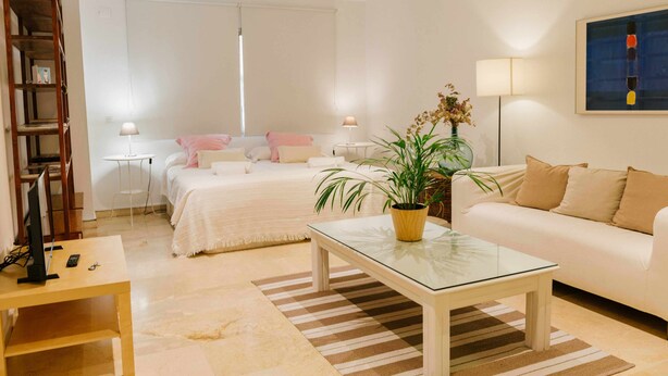 A modern hotel room with a bed, a sofa, a glass coffee table, a potted plant, and a painting on the wall.