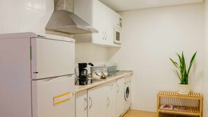A compact kitchen with a refrigerator, microwave, and washing machine. There's a small wooden shelf with a plant and magazines.