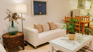 A living room with a white sofa, a glass coffee table, a wicker side table with a vase, and a painting on the wall.