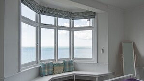 Een slaapkamer met een erker met uitzicht op zee, een witte bank met kussens, een bed met een paars deken en een spiegel aan de muur.