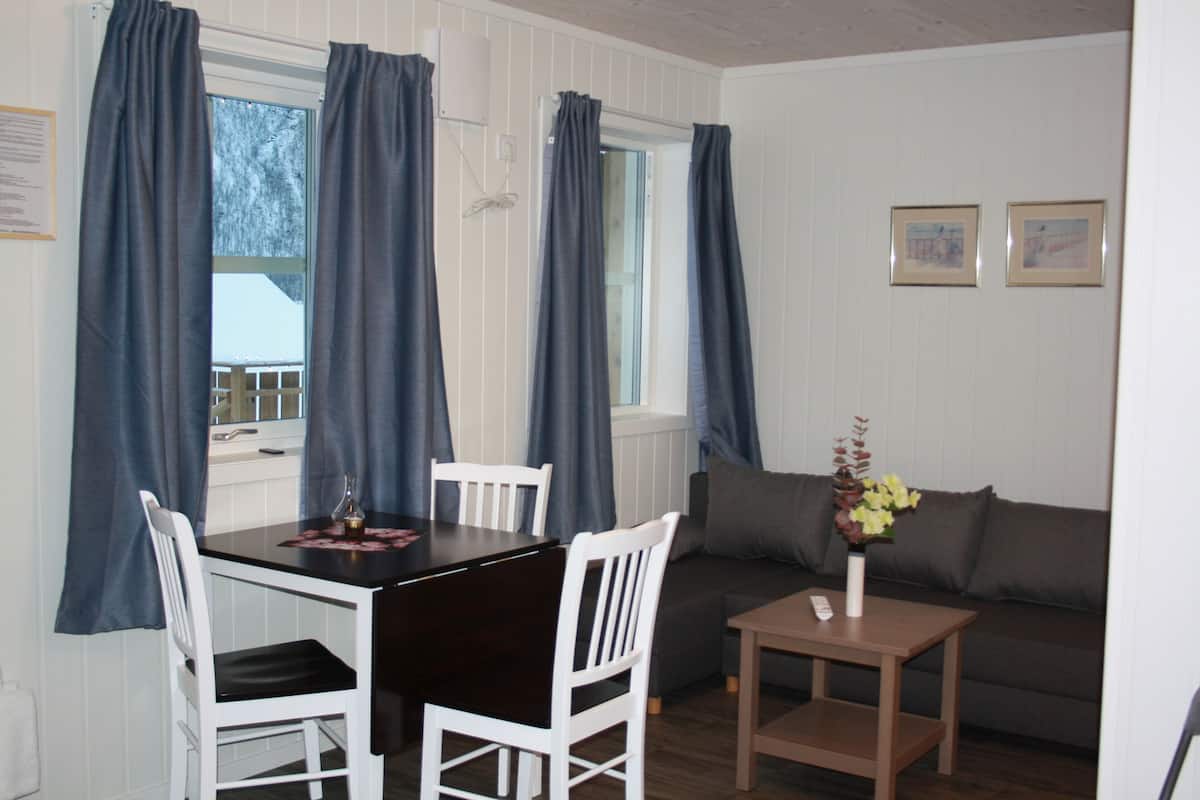 A room with a dining table, chairs, a sofa, a small table with a vase, and curtains on the windows.