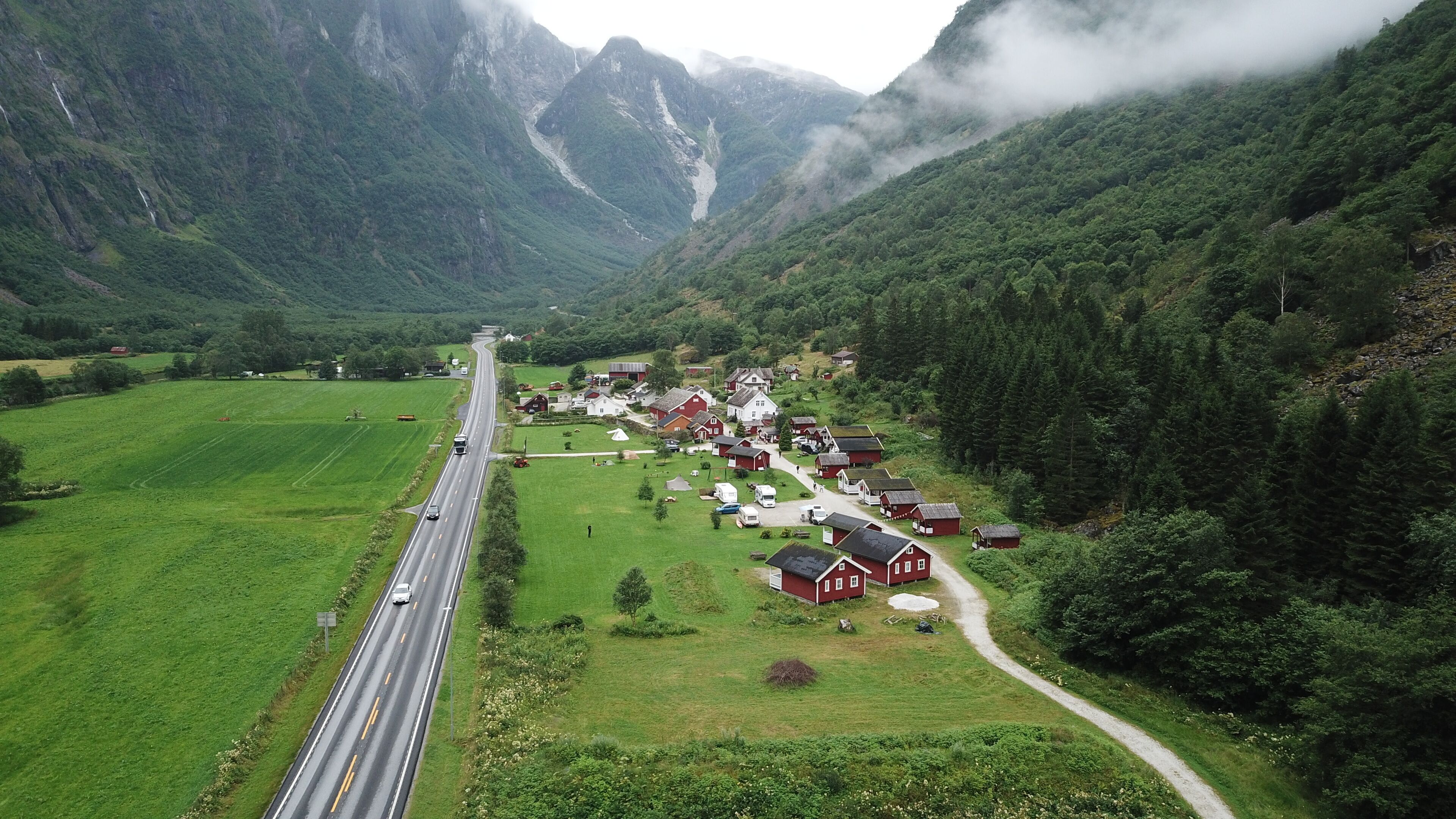 A rural road leading to a cluster of red houses surrounded by lush green fields and mountains.