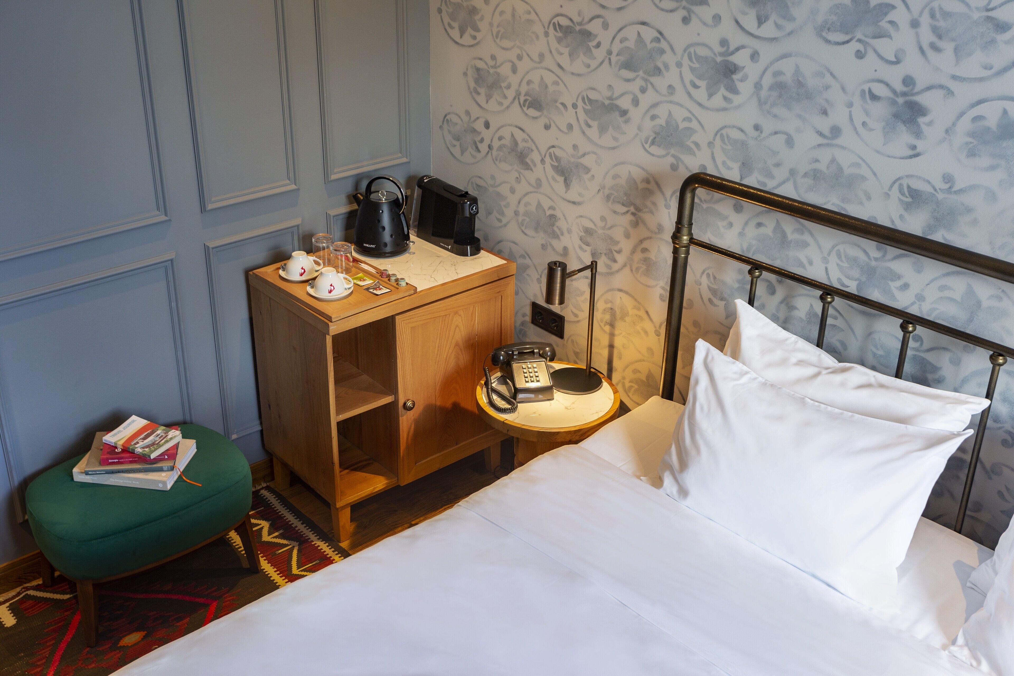 A hotel room with a bed, a wooden nightstand with a coffee maker, a telephone, and a green ottoman with books.