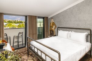 Hotellihuone, jossa on suuri sänky, tuoleilla varustettu parveke ja näkymä rakennuksiin ja viheralueille.