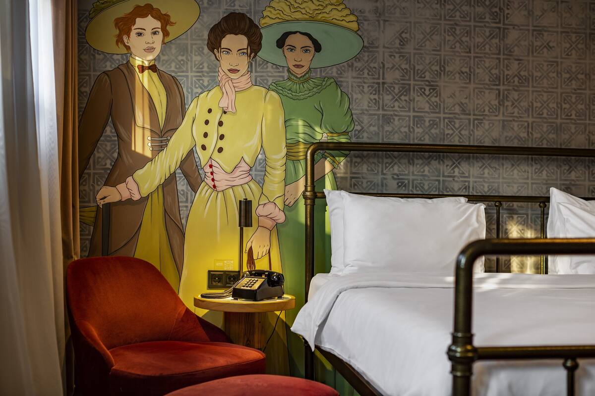 A hotel room with a bed, a red chair, a small table with a telephone, and a wall with a patterned wallpaper.