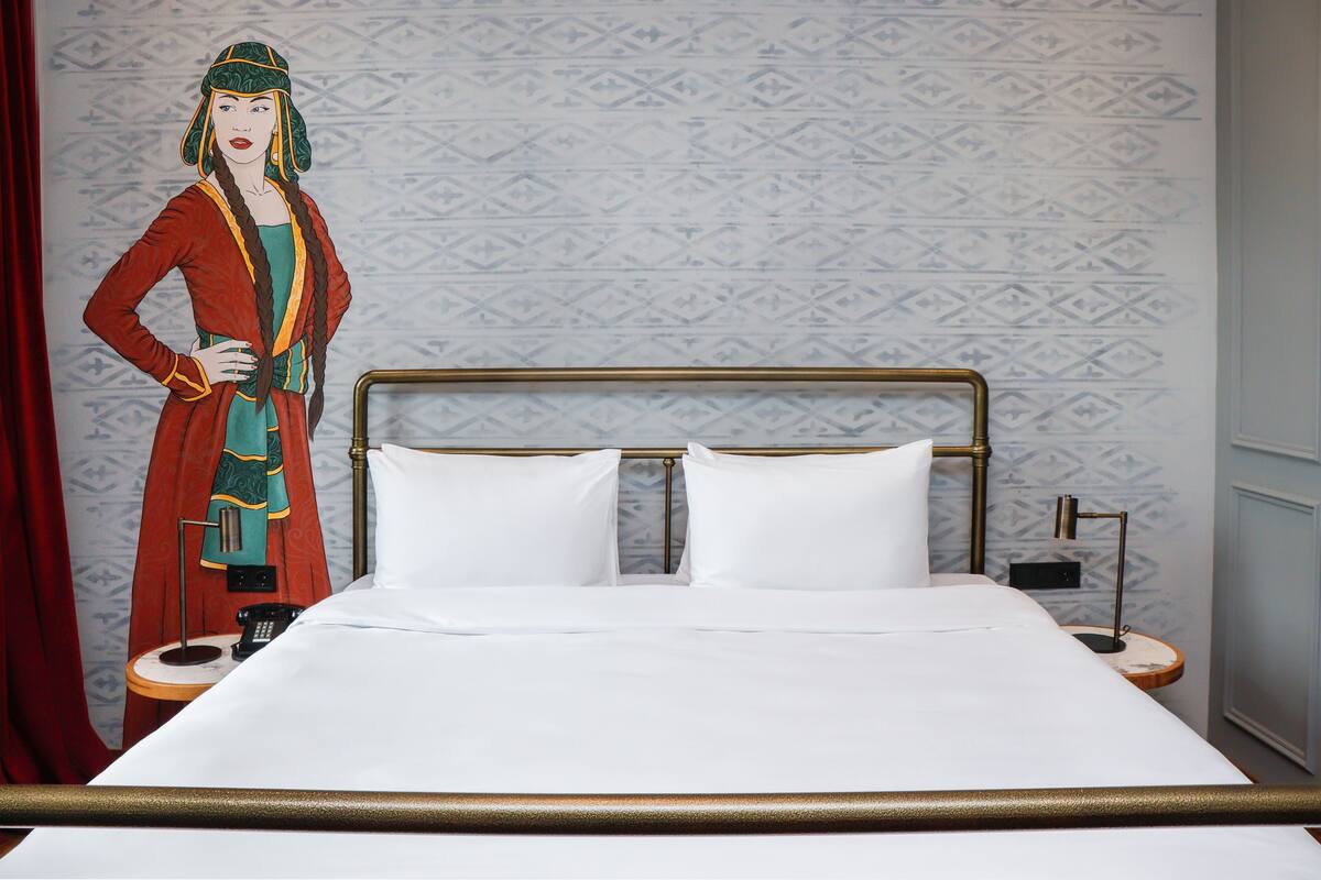 A hotel room with a bed, two bedside tables, a nightstand, and a wall with patterned wallpaper.
