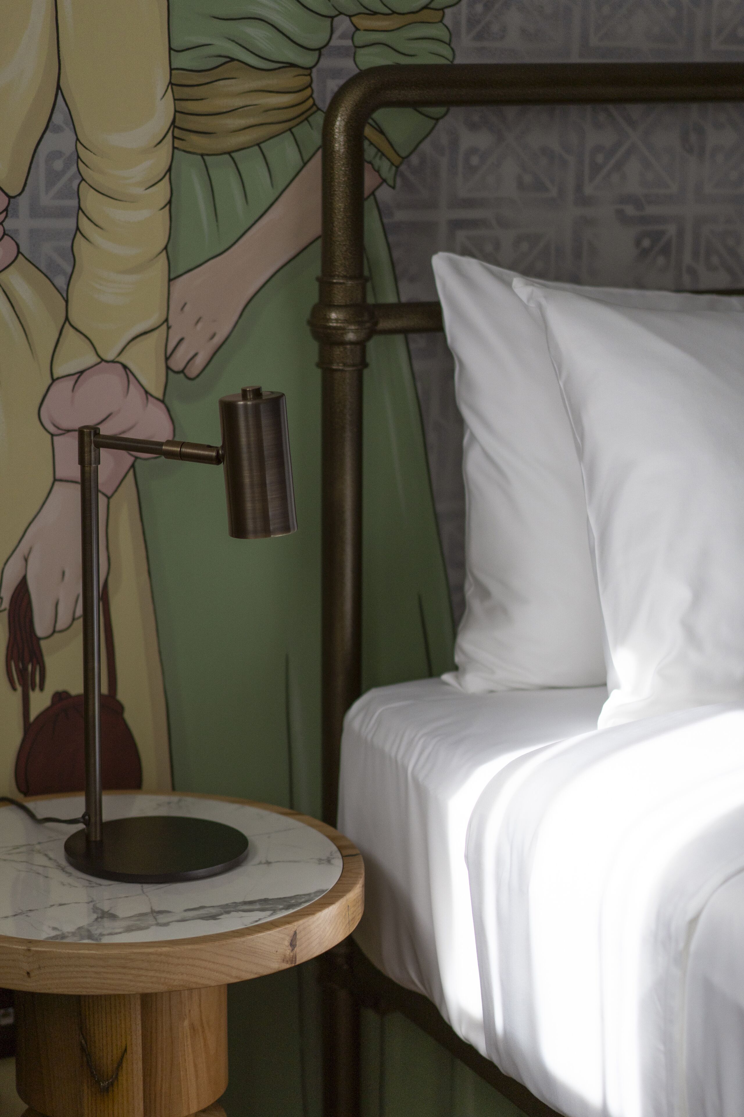 A bed with white bedding, a round wooden nightstand with a lamp, and a wall mural.