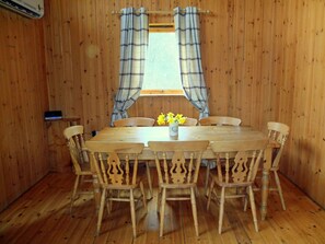 Una habitación con paneles de madera, una mesa redonda de madera y seis sillas. Hay una ventana con cortinas escocesas y un jarrón con flores amarillas sobre la mesa.