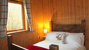 A wooden cabin bedroom with a bed, a window with curtains, a nightstand with a lamp, and a red throw blanket. - OAK is a charming, well equipped log cabin with hot tub and wood burning stove (Dalavich)