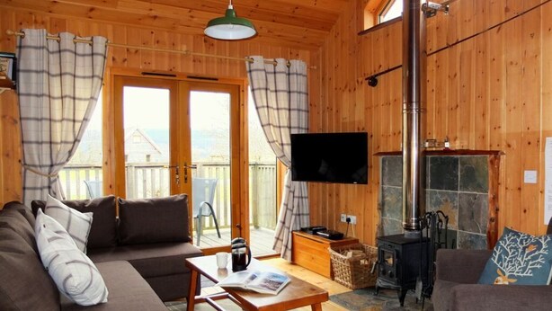 A cozy cabin interior with a sofa, a table with a magazine, a television, and a wood-burning stove. - OAK is a charming, well equipped log cabin with hot tub and wood burning stove (Dalavich)