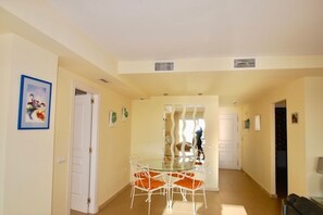 Un vaste espace dédié à la salle à manger, avec une table en verre et des chaises blanches, qui donne sur une cuisine et une porte.