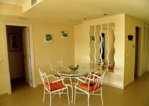 Un espace repas avec une table ronde en verre, entourée de chaises blanches dotées de coussins orange.