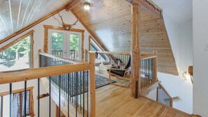Un interior de madera con una escalera que lleva a un balcón, una silla y una ventana con vistas a la vegetación.