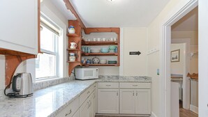 Une cuisine compacte avec des meubles blancs, un four à micro-ondes et une bouilloire posés sur le plan de travail. Une étagère en bois, située au-dessus du plan de travail, contient divers plats et un panneau indique la direction à suivre, vers la droite.