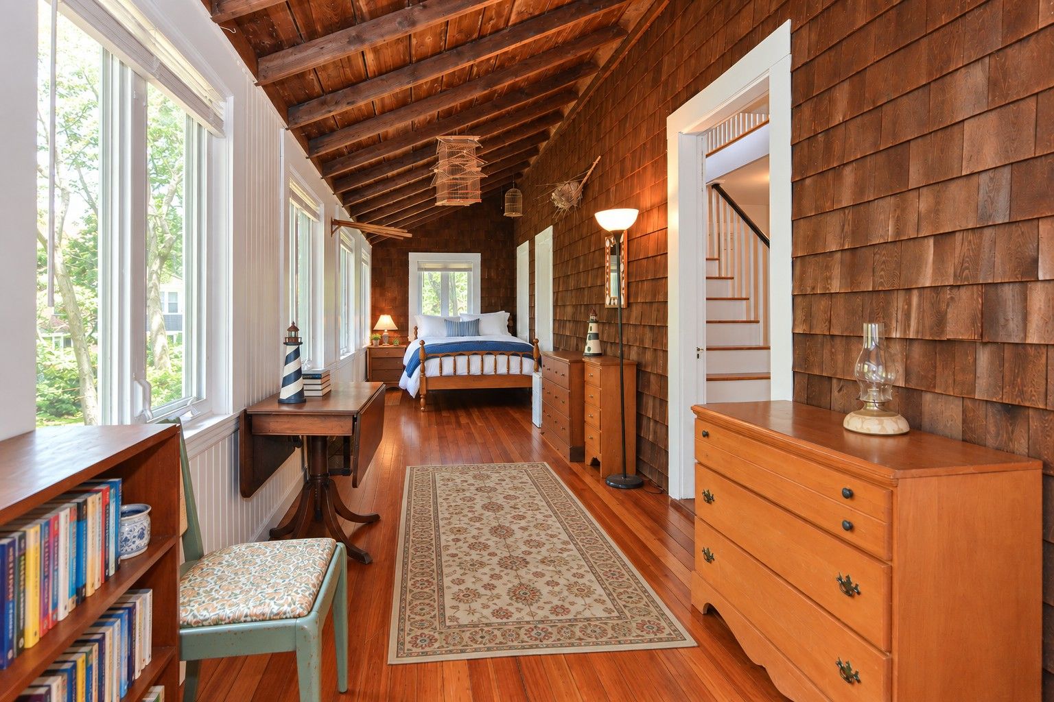 A bedroom with a wooden ceiling, a bed with a blue and white bedspread, a nightstand with a lamp, a dresser, and a bookshelf with books.